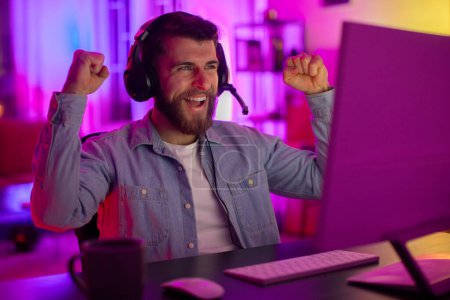Photo for A cheerful man wearing headphones with a mic celebrates with a triumphant gesture in a neon-lit room - Royalty Free Image