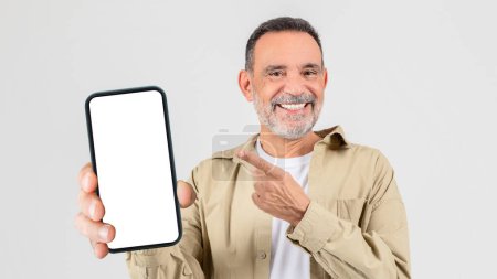 Téléchargez les photos : Un homme âgé joyeux avec du chaume dans une chemise décontractée tenant un smartphone avec un écran blanc vierge, le pointant avec son autre main - en image libre de droit