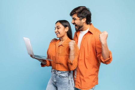 Two joyful Indian people in orange shirts celebrate with a laptop, conveying success or good news on blue