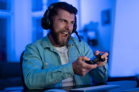 Photo for Happy bearded man with a gaming controller feels excitement during a game, blue light filling the room - Royalty Free Image