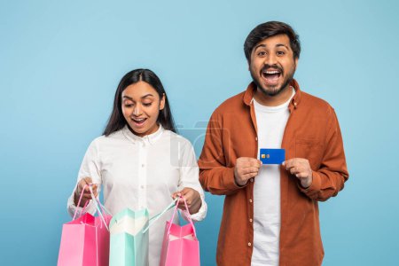 Aufgeregtes indisches Paar mit bunten Einkaufstüten und blauer Kreditkarte, das Konsumverhalten auf blauem Grund darstellt