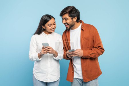 A happy Indian man and woman smile as they share content on a smartphone, conveying connection on blue