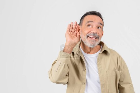 Foto de Anciano con un gesto de barba de mano a oreja que indica escuchar atentamente o dificultad auditiva sobre fondo claro - Imagen libre de derechos