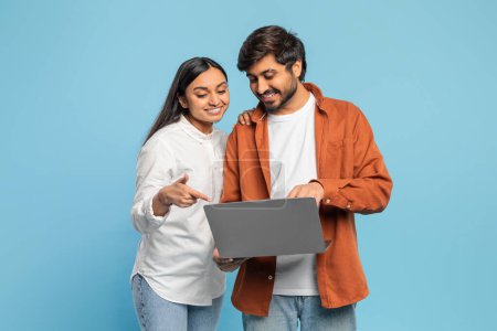Konzentriertes indisches Paar, das mit einem Laptop interagiert, vielleicht auf blauem Grund recherchiert oder online einkauft