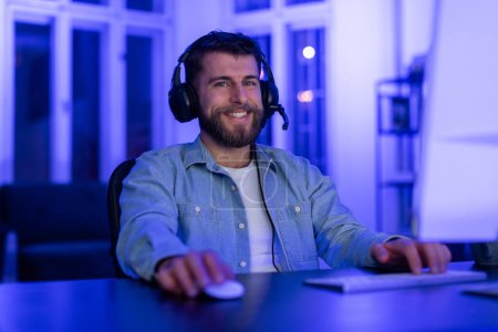 A man in a denim jacket with a headset sits at a desk with a computer at home, illuminated by blue light