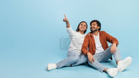 Alegre pareja india sentada en el suelo, señalando alegremente y mirando hacia otro lado de la cámara, copiando espacio