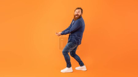 Téléchargez les photos : Prise de vue complète d'un homme aux cheveux roux positifs avec barbe tenant les mains ensemble avec les paumes levées, portant ou essayant d'attraper quelque chose d'invisible, debout au-dessus d'un mur de studio orange, panorama, espace de copie - en image libre de droit