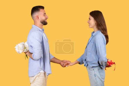 Téléchargez les photos : Un homme en chemise bleue tient un bouquet derrière son dos, tandis qu'il tient la main d'une femme en chemise de denim sur un fond jaune - en image libre de droit