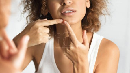 Foto de Afro Girl apretando espinilla en la cara frunciendo el ceño mirando en el espejo en el baño. Problemas de piel. Enfoque selectivo, Panorama - Imagen libre de derechos