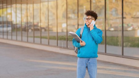 Jeune, brésilien enclin académique gars marche à l'extérieur tout en parlant au téléphone avec des ordinateurs portables à la main