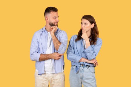 Photo for A man and woman stand facing each other with thoughtful expressions, hands on their chins against a yellow background - Royalty Free Image