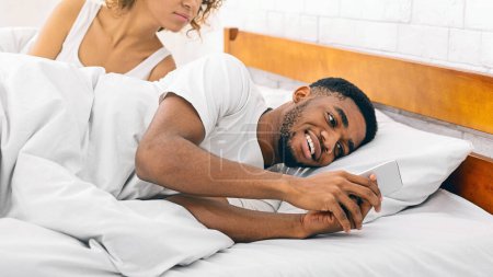 A candid moment of distrust, depicting African American man sneakily checking his phone beside his unaware partner in bed