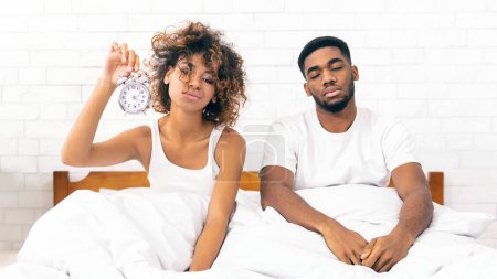 Téléchargez les photos : On devrait aller se coucher plus tôt. Couple afro-américain endormi assis au lit avec réveil, ne peut se réveiller le matin, espace de copie - en image libre de droit