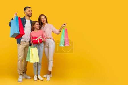 Téléchargez les photos : Une famille joyeuse présentant un cadeau et tenant des sacs à provisions, présentant un style de vie de consommateur sur une toile de fond jaune - en image libre de droit