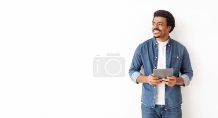 Foto de Relajado hombre negro en traje de mezclilla sosteniendo tableta digital y mirando a un lado, guapo joven afroamericano macho de pie contra la pared blanca en una habitación espaciosa, mirando el espacio de copia - Imagen libre de derechos