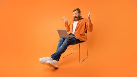 Photo for Redhaired bearded man looks at laptop and shouting with shocked expression, experiencing issue during websurfing and online work, sitting with computer in chair over orange wall. Panorama - Royalty Free Image