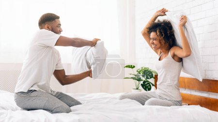 Energetisches afrikanisch-amerikanisches Paar bei einer fröhlichen Kissenschlacht in einem sonnigen, modernen Schlafzimmer-Interieur