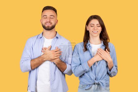 A man and woman stand with eyes closed, hands on their hearts, expressing thankfulness and sincerity on a yellow background