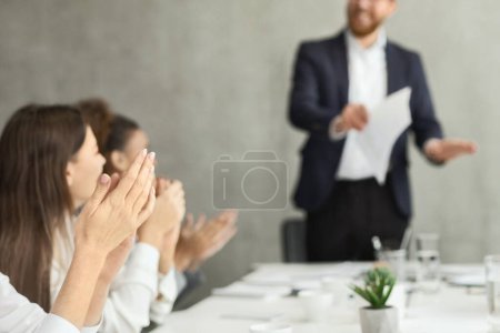 Photo for A blurred audience applauds a confident businessman presenting documents at a modern conference table - Royalty Free Image