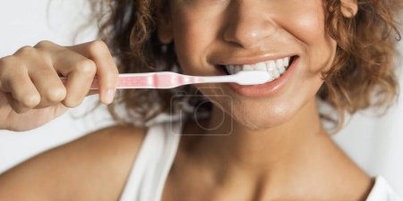 Foto de Mujer afro joven sonriente con cepillo de dientes limpiando los dientes en el baño. Concepto diario de cuidado dental - Imagen libre de derechos