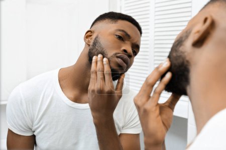 Hombre afroamericano toca su cara mientras examina de cerca su piel, probablemente en busca de manchas o imperfecciones