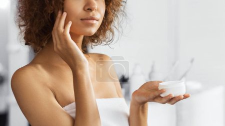 Belle fille afro-américaine appliquant de la crème sur son visage, debout dans la salle de bain
.