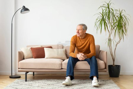 Hombre mayor sentado cómodamente en un sofá en medio de almohadas de tonos cálidos, con una planta en maceta en el fondo