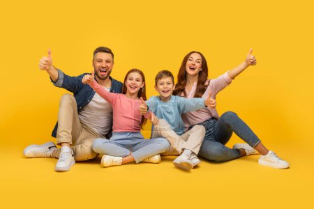 Foto de Una familia alegre de cuatro con dos hijos y padres dando pulgares hacia arriba, todos sonriendo en un vibrante telón de fondo amarillo - Imagen libre de derechos