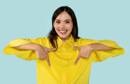 Photo for Cheerful brunette in vibrant yellow shirt playfully pointing downwards with both hands, positive young woman against trendy blue background. Promotions and attention-grabbing ads concept - Royalty Free Image