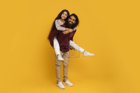Téléchargez les photos : Un homme joyeux en chemise bordeaux tient une femme sur le côté, souriant tous les deux sur un fond jaune vif - en image libre de droit