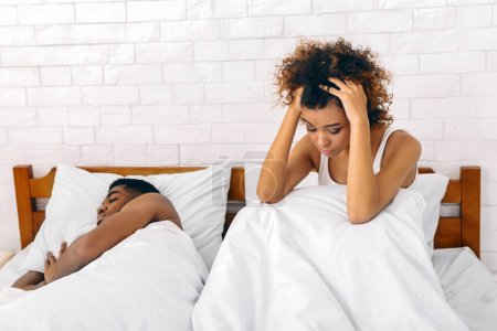 Foto de Afectada mujer afroamericana sostiene la cabeza en las manos junto a un hombre dormido, que representa la tensión emocional en una relación - Imagen libre de derechos