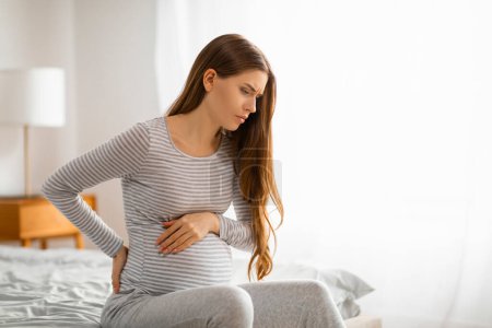 A pregnant woman shows signs of discomfort or cramps, sitting on the bed with a worried expression