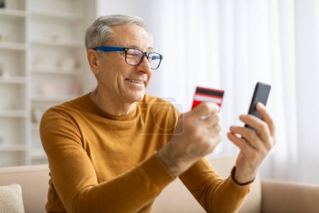 Téléchargez les photos : Homme âgé portant des lunettes évaluant les informations financières sur sa carte de crédit et son smartphone à la maison - en image libre de droit