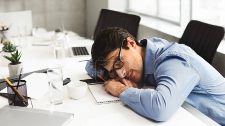 Photo for A tired man worker in a blue shirt is slumped over his desk next to a laptop and a cup of coffee, illustrating fatigue and overwork in the workplace - Royalty Free Image