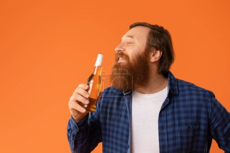 Téléchargez les photos : Heureux redhaired barbu homme dans occasionnel aime la bière, posant avec bouteille et consommer boisson alcoolisée préférée sur fond de studio orange, portrait shot, copier l'espace - en image libre de droit