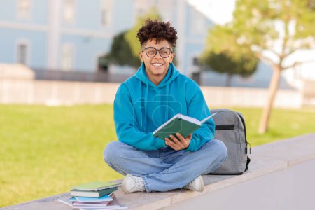 A smiling young brazilian student with glasses is sitting outside with a book in hand, in a relaxed posture with a backpack and notebooks nearby