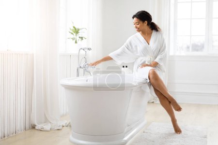 Téléchargez les photos : Jeune femme afro-américaine assise sur une baignoire blanche autoportante dans un cadre de salle de bain moderne et lumineux - en image libre de droit
