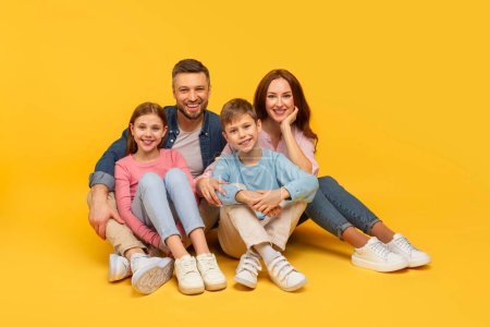 Téléchargez les photos : Une famille heureuse avec deux enfants assis près du sol, tous souriants en tenue décontractée sur fond jaune - en image libre de droit