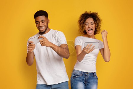 Foto de Un hombre y una mujer afroamericanos alegres están jugando en sus teléfonos inteligentes contra un fondo amarillo vivo, ambos mostrando expresiones de emoción y alegría - Imagen libre de derechos