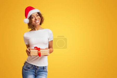 Téléchargez les photos : Heureuse fille afro-américaine au chapeau de Père Noël tenant cadeau de Noël, regardant la caméra et souriant, fond de studio orange - en image libre de droit