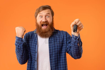 New Car. Emotional Bearded Redhaired Man Showing Automobile Key And Gesturing Yes, Shouting In Excitement, Celebrating Auto Purchase Or Loan Against Orange Studio Background