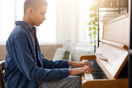 Téléchargez les photos : Un jeune homme habillé de façon décontractée pratique sur un piano droit dans une pièce confortable et ensoleillée, les mains en mouvement - en image libre de droit