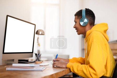 Photo for Engaged black teenage student guy using headphones while studying at desk with computer with blank monitor, writing at notebook in sunny room - Royalty Free Image