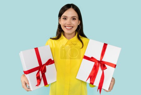 Photo for Radiant young brunette woman in yellow blouse joyfully presenting white gift boxes with bold red ribbons, against light blue backdrop. Perfect capture for festive occasions and gifting. - Royalty Free Image