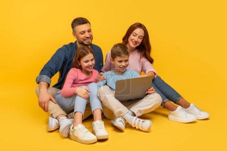 Foto de Familia sonriente sentada en el suelo interactuando con un portátil, comprometida y feliz, sobre un vibrante telón de fondo amarillo - Imagen libre de derechos