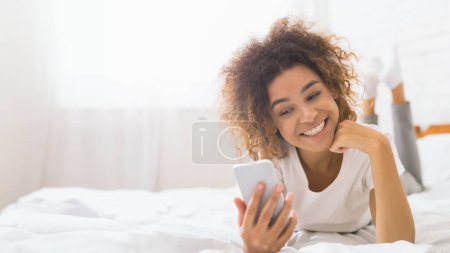 Téléchargez les photos : Gros plan d'une jeune femme afro-américaine joyeuse aux cheveux bouclés utilisant son téléphone au lit, envoyant éventuellement des SMS ou parcourant les médias sociaux - en image libre de droit
