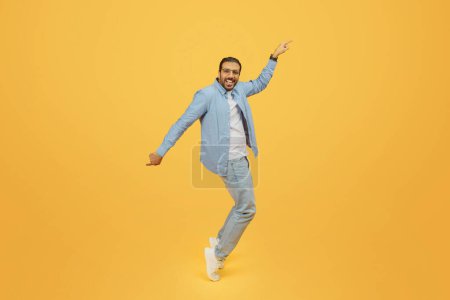 An exuberant bearded indian man dances with joy, his arm outstretched pointing upwards, wearing a denim shirt and glasses, against a yellow backdrop