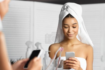 Téléchargez les photos : Toujours en contact. Femme noire avec serviette sur la tête textos sur Smartphone debout dans la salle de bain. Panorama - en image libre de droit