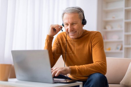 Un anciano feliz se dedica a una conversación usando un auricular con un micrófono, sentado frente a un portátil