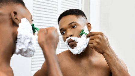 Afroamerikaner rasiert sich sein Gesicht vorsichtig mit einem Rasiermesser, während er in den Badezimmerspiegel schaut, wobei Rasierschaum sein Gesicht bedeckt